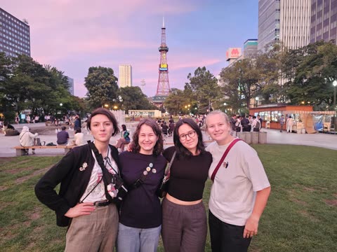 Four women stand together smiling in a city park at dusk, with the Sapporo TV Tower visible in the background.