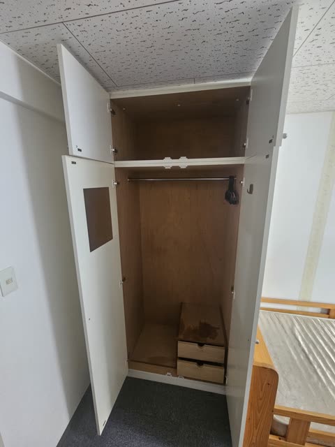 An empty white wardrobe with its doors open stands in a carpeted room next to a wooden bed frame.