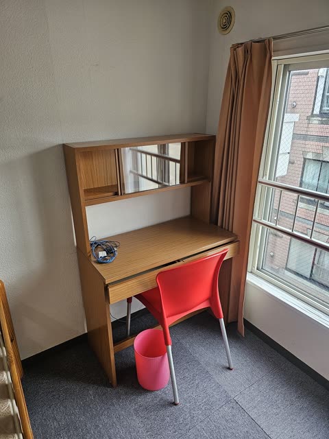 A wooden desk with a hutch and a red chair are set in a room's corner next to a window with brown curtains.