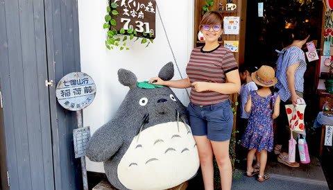 A person in a striped shirt and glasses stands with their hand on a large, grey plush Totoro figure outside a shop entrance.