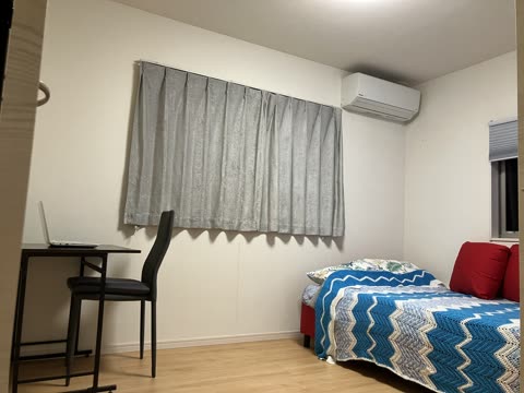 A bedroom with a wood floor contains a bed with a blue crocheted blanket and a small desk with a laptop.