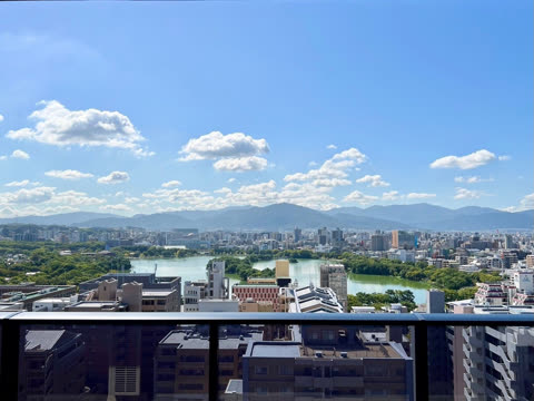 A cityscape surrounding a green lake is viewed from a high balcony, with a mountain range visible in the distance.