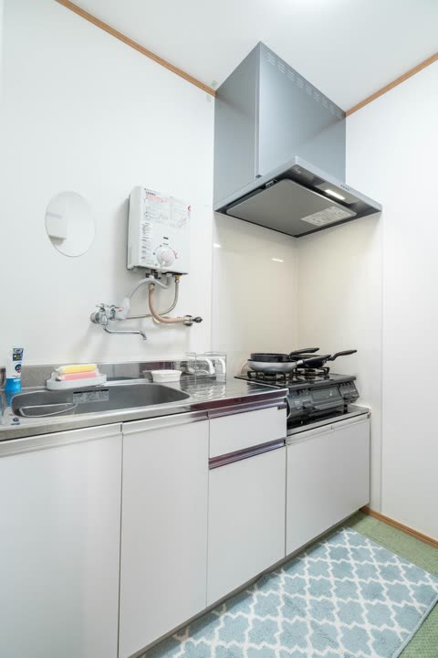 A compact kitchen with a stainless steel sink, a gas stovetop with pans, and a silver range hood against a white wall.