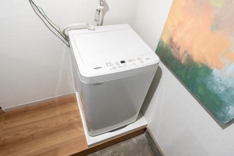 A white washing machine sits on a raised platform in a corner next to a large, colorful painting.