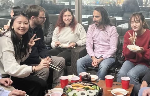 A group of people sits around a low table, smiling and eating food cooked on a tabletop grill with chopsticks.