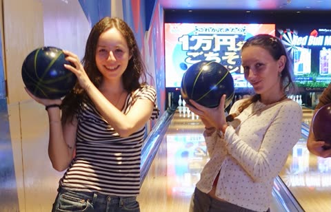 Two women holding bowling balls smile for the camera while standing next to a lane in a bowling alley.