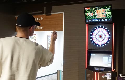 A person in a white sweatshirt and black cap aims a dart at an illuminated electronic dartboard machine indoors.