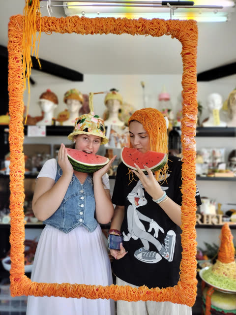Two women hold watermelon slices to their mouths while posing behind a noodle-wrapped frame in a food-themed hat shop.