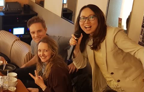 Three people are in a room, where one woman holds a microphone, another makes a peace sign, and a man smiles.