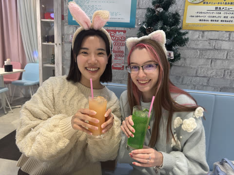 Two women in bunny and cat ear headbands smile while holding colorful drinks in a cafe booth.
