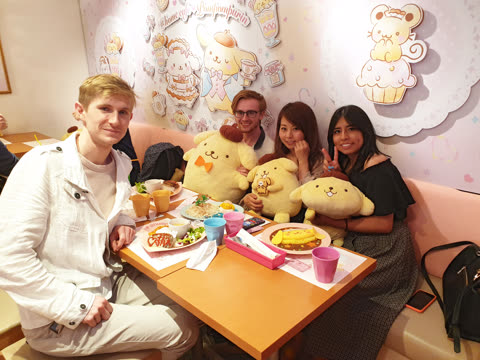 Four people sit at a cafe table with Pompompurin plushies, eating meals.