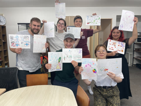 A group of seven adults smiles while holding up their hand-drawn illustrations and comics in an office setting.