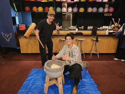 Two men smile, one holding a wooden mallet and one kneeling by a stone mortar with white dough, inside a restaurant.