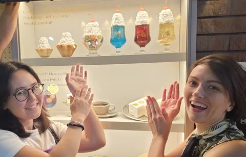 Two women smile with their hands raised in front of a display case of Japanese plastic food models.