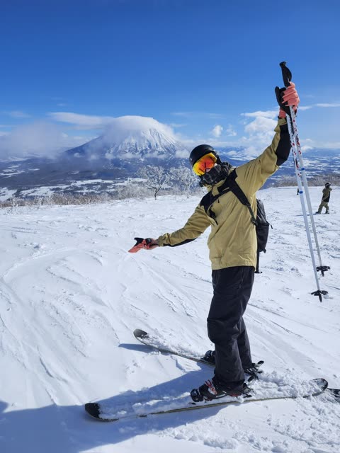 A skier in a tan jacket raises a ski pole while standing on a snowy mountain with another large peak in the background.