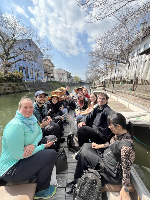 A group of people sits together on a long wooden boat in a canal lined with houses and trees.