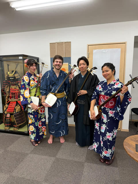 Four people in kimonos and yukatas stand holding shamisen instruments in a room with a samurai armor display.