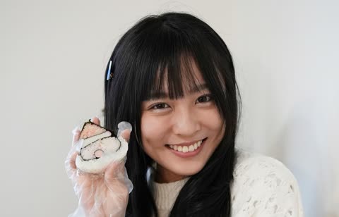 A smiling woman with black hair holds up a piece of Santa Claus-shaped onigiri against a plain, light-colored wall.