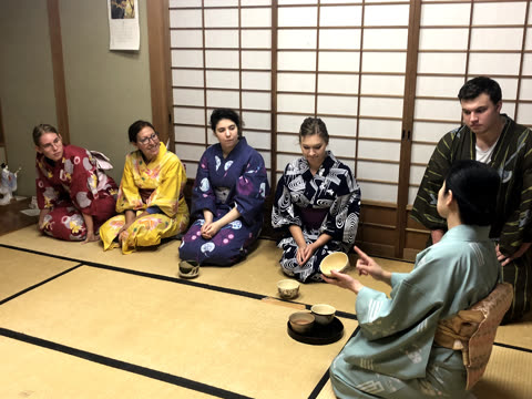 A woman in a kimono demonstrates with a tea bowl for a group of people sitting on a tatami mat floor.