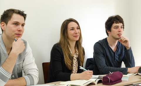 Three people sit at a desk in a classroom, looking forward with notebooks and pens.