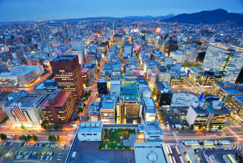 An aerial view of a dense cityscape at dusk, with illuminated buildings and traffic light trails leading toward distant mountains.