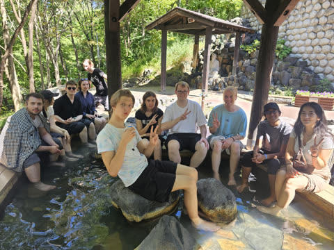 A group of people sits on wooden benches with their feet in a stone-lined pool of water in a sunlit garden.