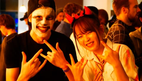 Two people in costumes, one with face paint and another with devil horns, make rock-on hand gestures in a crowded room.