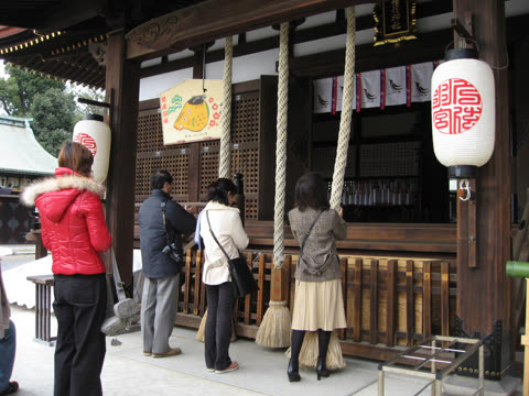Four people stand with their backs to the viewer, holding thick ropes in front of a traditional wooden Japanese shrine.