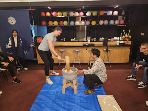 A man raises a wooden mallet over a stone mortar as another man kneels beside him inside a room with paper lanterns.