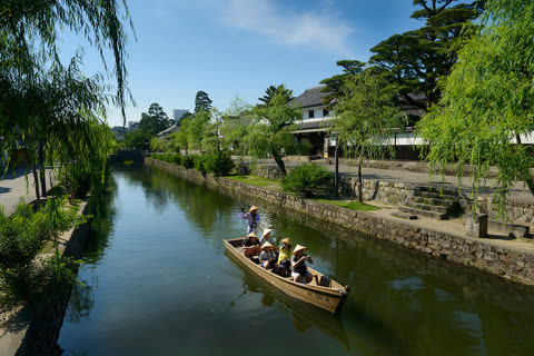 A person poles a wooden boat with passengers wearing conical hats down a canal lined with willow trees and buildings.