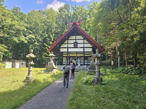 People walk along a path lined with stone lanterns toward a traditional Japanese building in a forest.