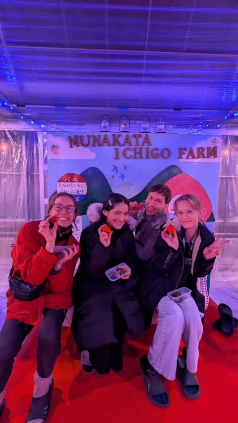 Four people in winter coats smile and hold up strawberries while sitting in front of a Munakata Ichigo Farm backdrop.