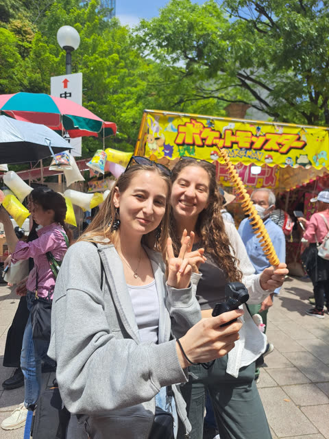 Two women pose for a photo at an outdoor street fair, one holding a camera and the other holding a spiral-cut potato.