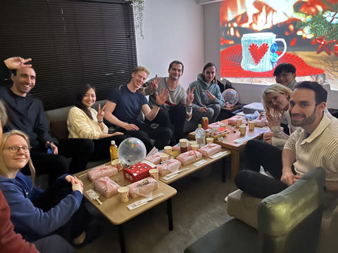 A group of people sit on couches around low tables, smiling and making hand gestures for a photo in a living room.