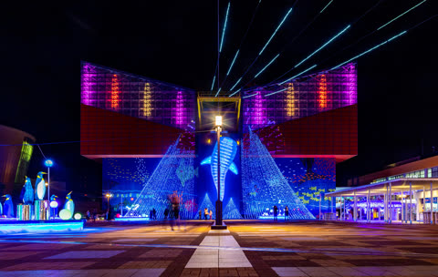A modern building and plaza are illuminated at night with large, colorful light displays of a whale shark, penguins, and trees.