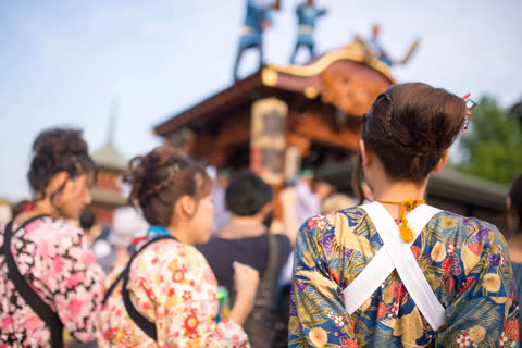 A person viewed from behind in a floral Japanese garment watches people standing on an ornate wooden festival float.