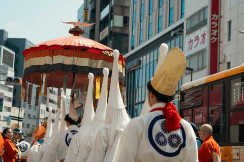People in traditional white robes and tall hats carry a large red parasol during a procession on a modern city street.