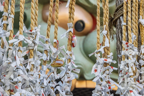 White paper fortunes and small charms are tied to thick, braided ropes hanging in front of a large, out-of-focus statue.