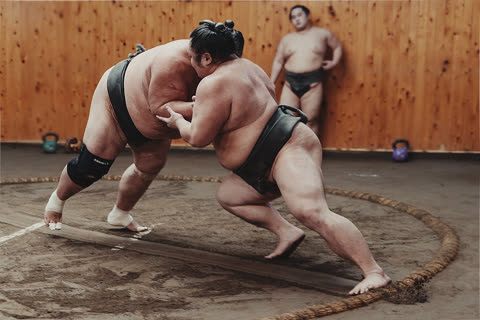 Two sumo wrestlers grapple inside a dirt ring as a third wrestler stands against a wood-paneled wall.