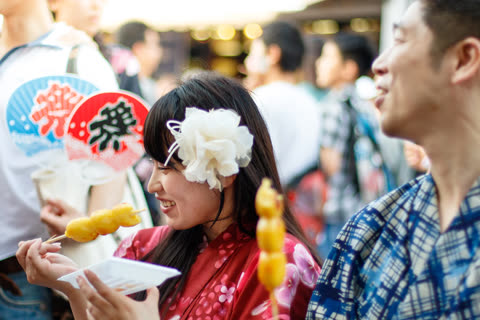 A woman in a red floral yukata with a white flower in her hair holds skewered food and smiles at a man at a festival.