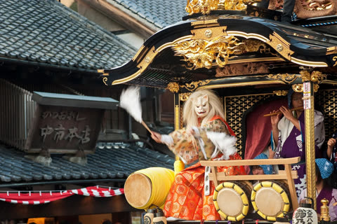 A person in a white mask and traditional costume sits on an ornate parade float, waving a white feathered wand.