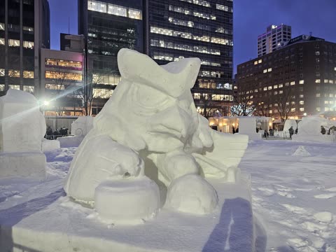 Snow sculpture of a bird-like figure playing curling, in a city square at night.