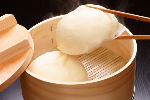 A pair of chopsticks lifts a steaming bun from an open wooden steamer basket.