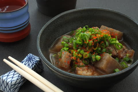 A bowl of stew with meat and konjac, garnished with sliced green onions and red spice, sits next to chopsticks on a dark surface.