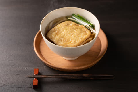 A steaming bowl of udon noodle soup with fried tofu and green onions sits on a wooden tray next to chopsticks on a dark table.