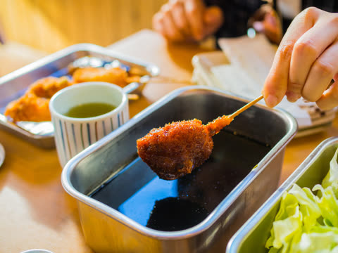 A hand dips a deep-fried, breaded skewer of food into a metal container of dark sauce on a wooden table.