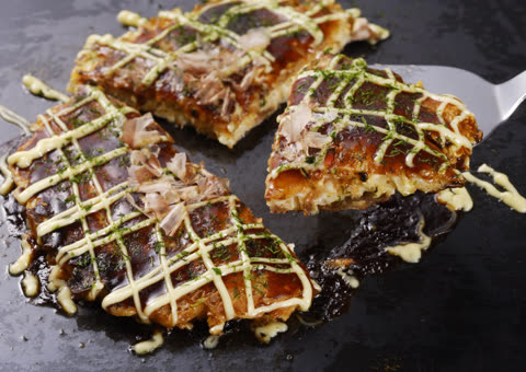 A metal spatula lifts a slice of okonomiyaki topped with sauce, mayonnaise, and bonito flakes from a teppanyaki grill.