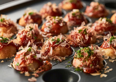 Round, fried dough balls are topped with sauce, thin flakes, and green herbs in a dimpled baking pan.