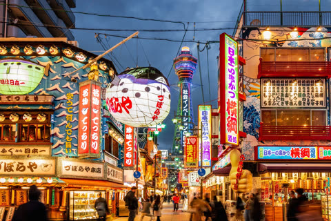A large, illuminated pufferfish lantern hangs over a bustling city street where people walk past glowing neon signs at dusk.