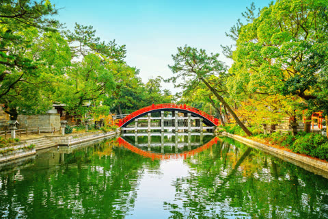 A red arched bridge spans a calm canal, its reflection mirrored in the water surrounded by lush green trees.